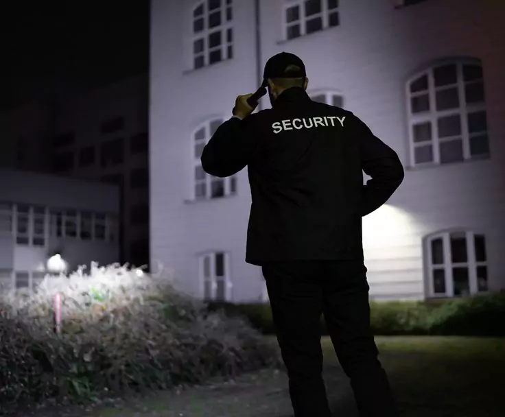 Agent de Sécurité Agadir en veste 'SECURITY' parle sur talkie-walkie la nuit près d’un bâtiment éclairé et d’arbustes.