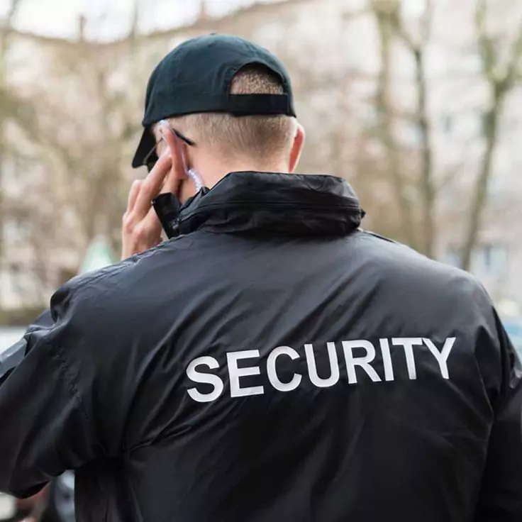 Agent de Sécurité Agadir en veste noire 'SECURITY', avec casquette, parle sur talkie-walkie en mission de gardiennage, fond flou.