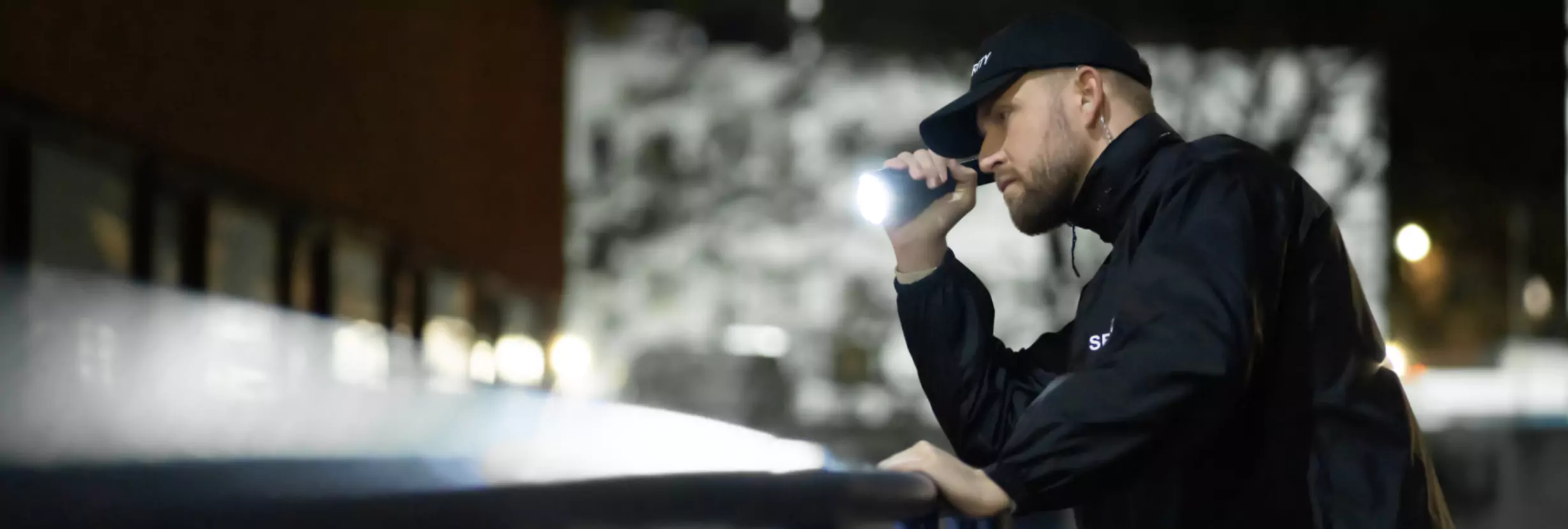 Agent de Sécurité Agadir en veste noire utilise une lampe torche et un talkie-walkie près d’un bâtiment la nuit.